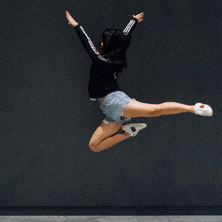 Eine Frau springt in einer Tanzpose vor einer schwarzen Wand hoch.