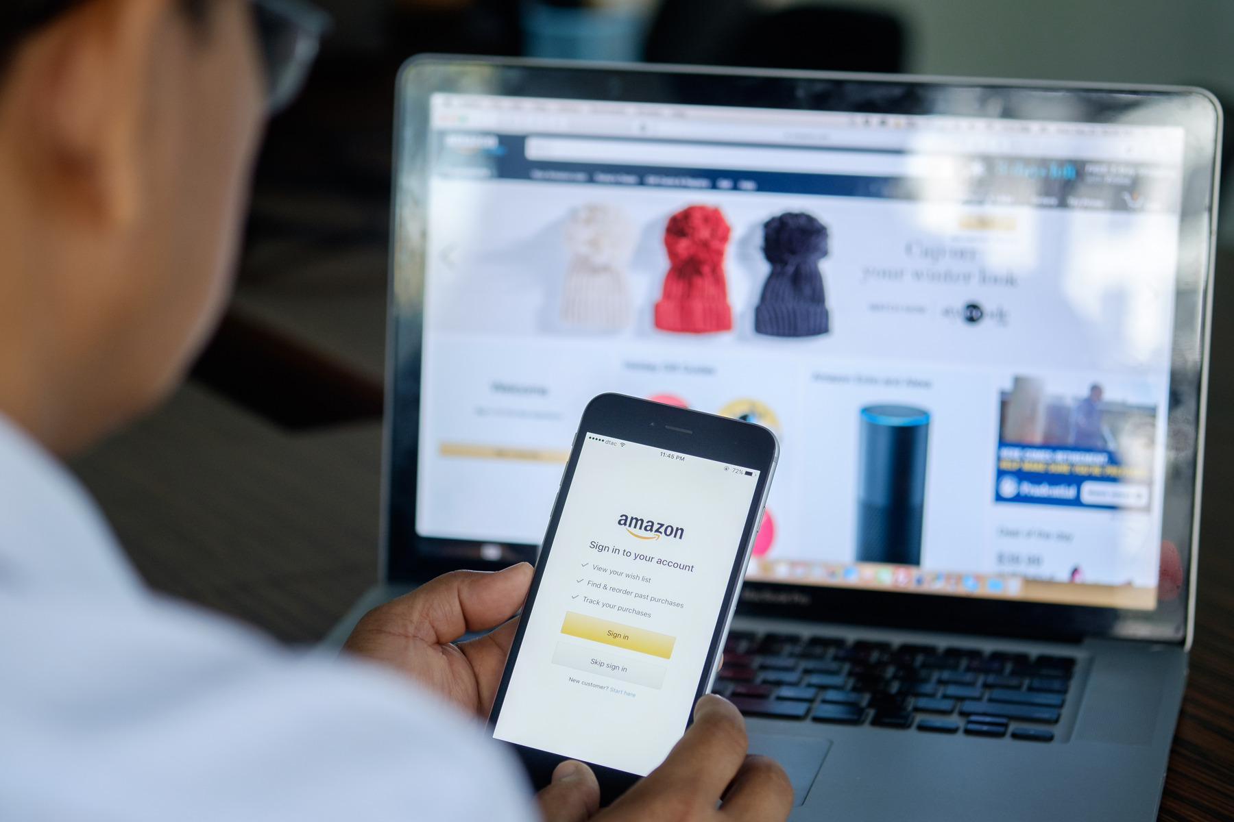 shutterstock_540533869_Premier_redaktionell.jpg_ST-CC Ein Mann hält ein Smartphone, auf dem die App Amazon zu sehen ist. Im Hintergrund ist ein Laptop, auf dem die Webseite von Amazon zu sehen ist.