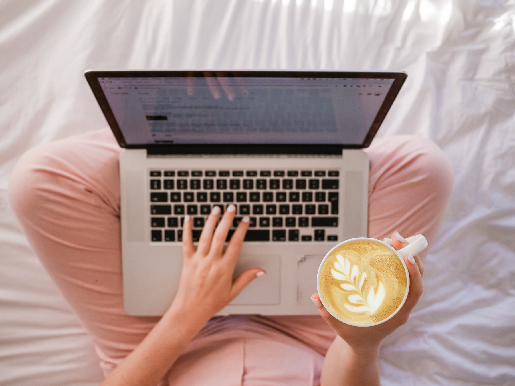 Das Foto zeigt eine Person, die im Bett arbeitet, mit Blick von oben. Sie sitzt im Schneidersitz auf dem Bett und hat den Laptop auf ihren Beinen abgestellt. In der rechten Hand hält sie einen mit aufwendiger Schaumkunst gestalteten Kaffee, der beste Barista-Skills vermuten lässt.