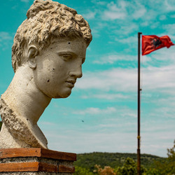 : Zu sehen sind der Kopf einer antiken Statue vor einer mediterranen Hügellandschaft im Nationalpark Butrint. Im Hintergrund weht die albanische Flagge.