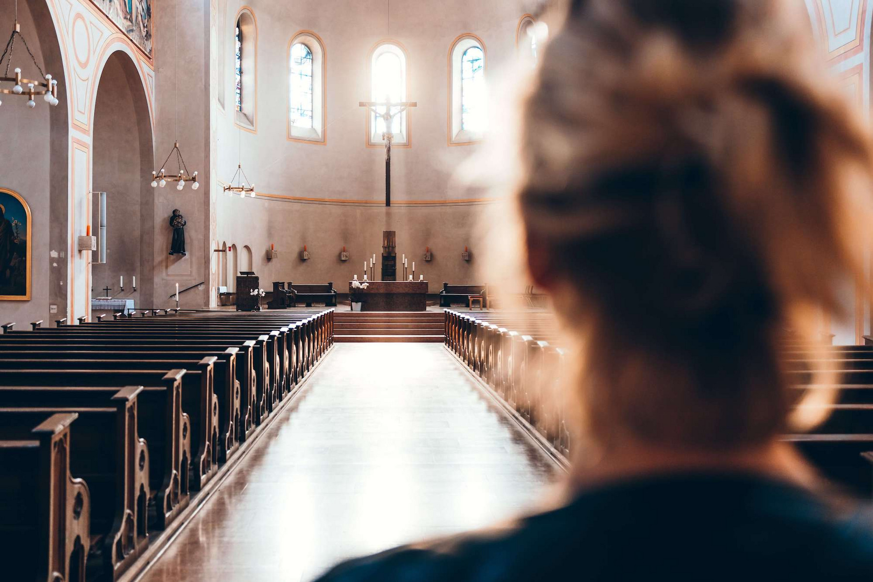 Eine Frau von hinten in einer leeren Kirche, sie blickt auf den sonnenbeschienenen Altar mit Jesus am Kreuz, der am Ende einer langen Bankreihe steht.