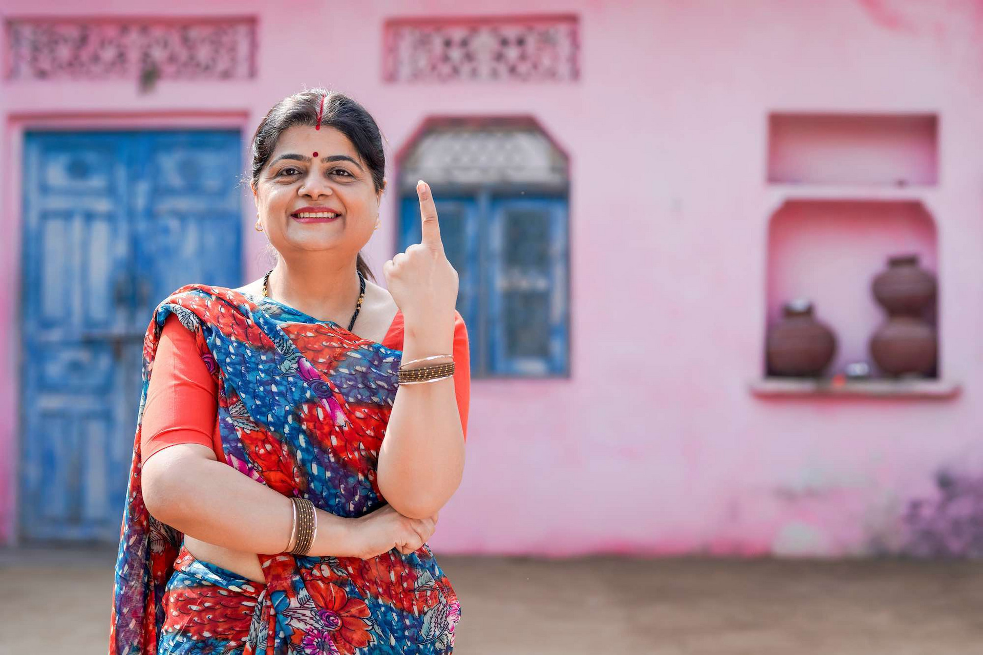 Eine indische Frau mit erhobenem Zeigefinger steht vor einem rosa Gebäude; in Indien wählt man mit dem Fingerabdruck des Zeigefingers