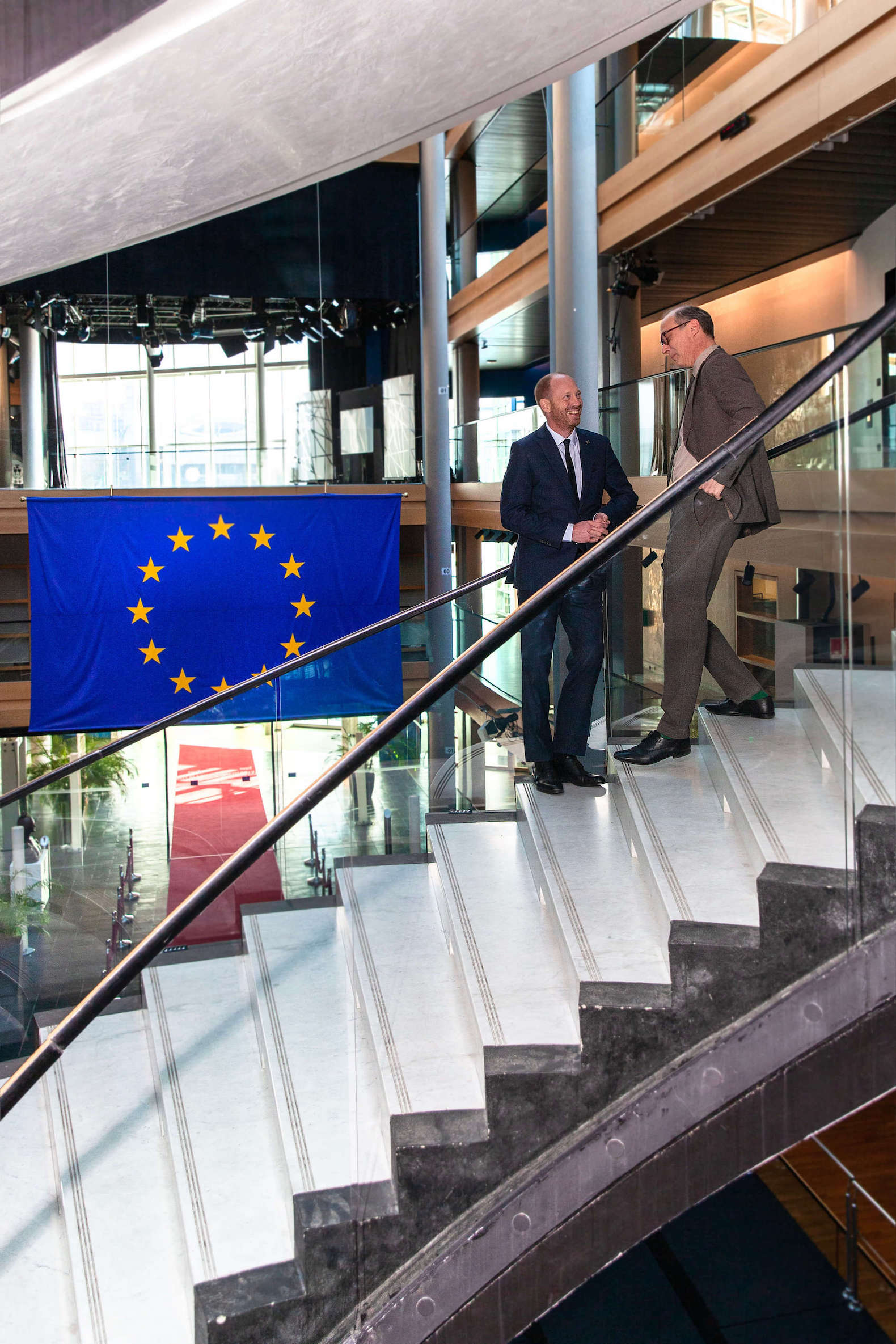 Zwei Männer unterhalten sich auf einer Treppe, im Hintergrund hängt eine EU-Flagge