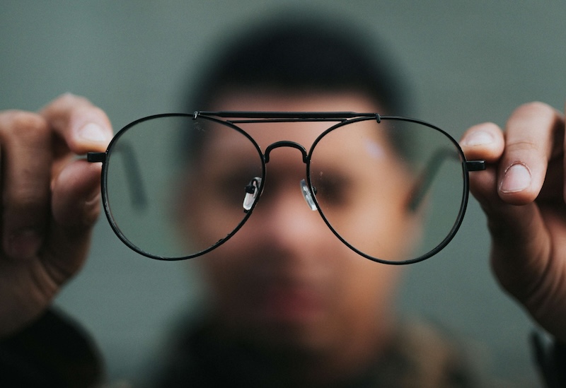 Ein Mann hält mit beiden Händen eine Brille in die Kamera, er selbst ist nur verschwommen im Hintergrund zu sehen.
