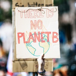 Ein handgemaltes Protestschild mit der Aufschrift "THERE IS NO PLANET B" und einer gezeichneten Weltkugel wird bei einer Demonstration hochgehalten. Im unscharfen Hintergrund sind weitere Menschen und Gebäude zu sehen.