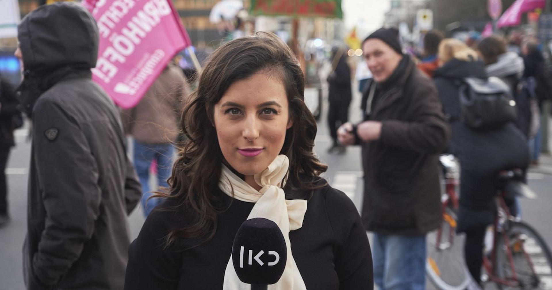 Eine Frau mit einem Mikrofon in der Hand steht mitten in einer Demonstration und schaut in die Kamera.