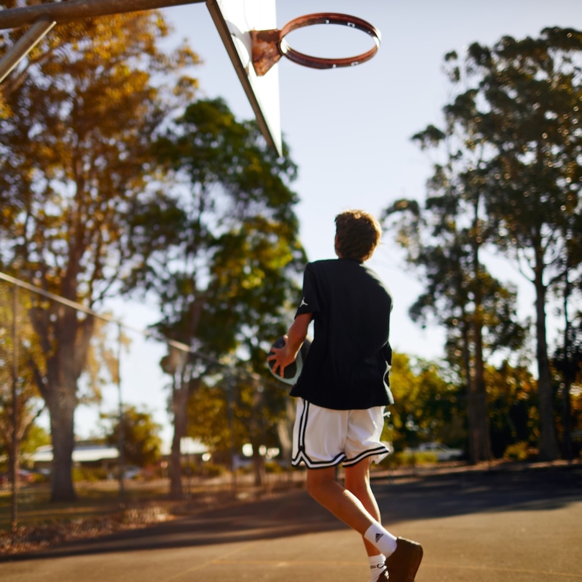 Ein Jugendlicher läuft auf einem Basketballplatz im Freien auf einen Korb zu. Im Hintergrund sind Bäume und warmes Sonnenlicht zu sehen. Die Szene wirkt ruhig und sommerlich.