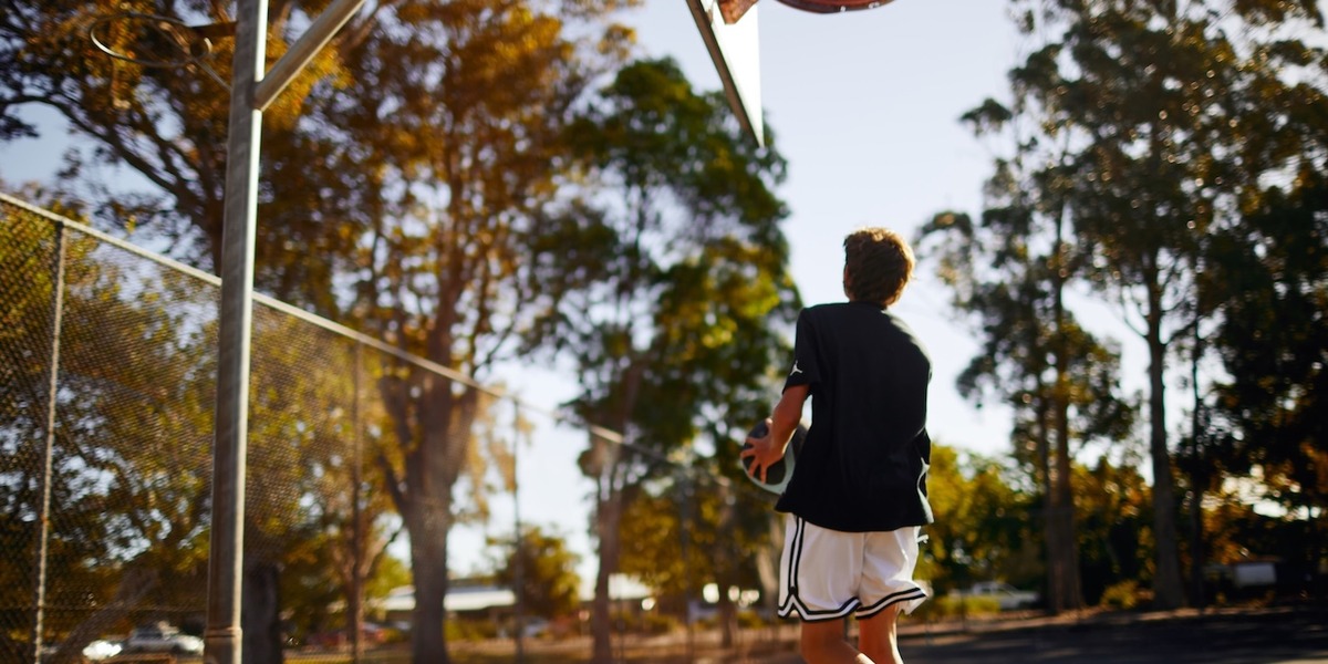 Ein Jugendlicher läuft auf einem Basketballplatz im Freien auf einen Korb zu. Im Hintergrund sind Bäume und warmes Sonnenlicht zu sehen. Die Szene wirkt ruhig und sommerlich.