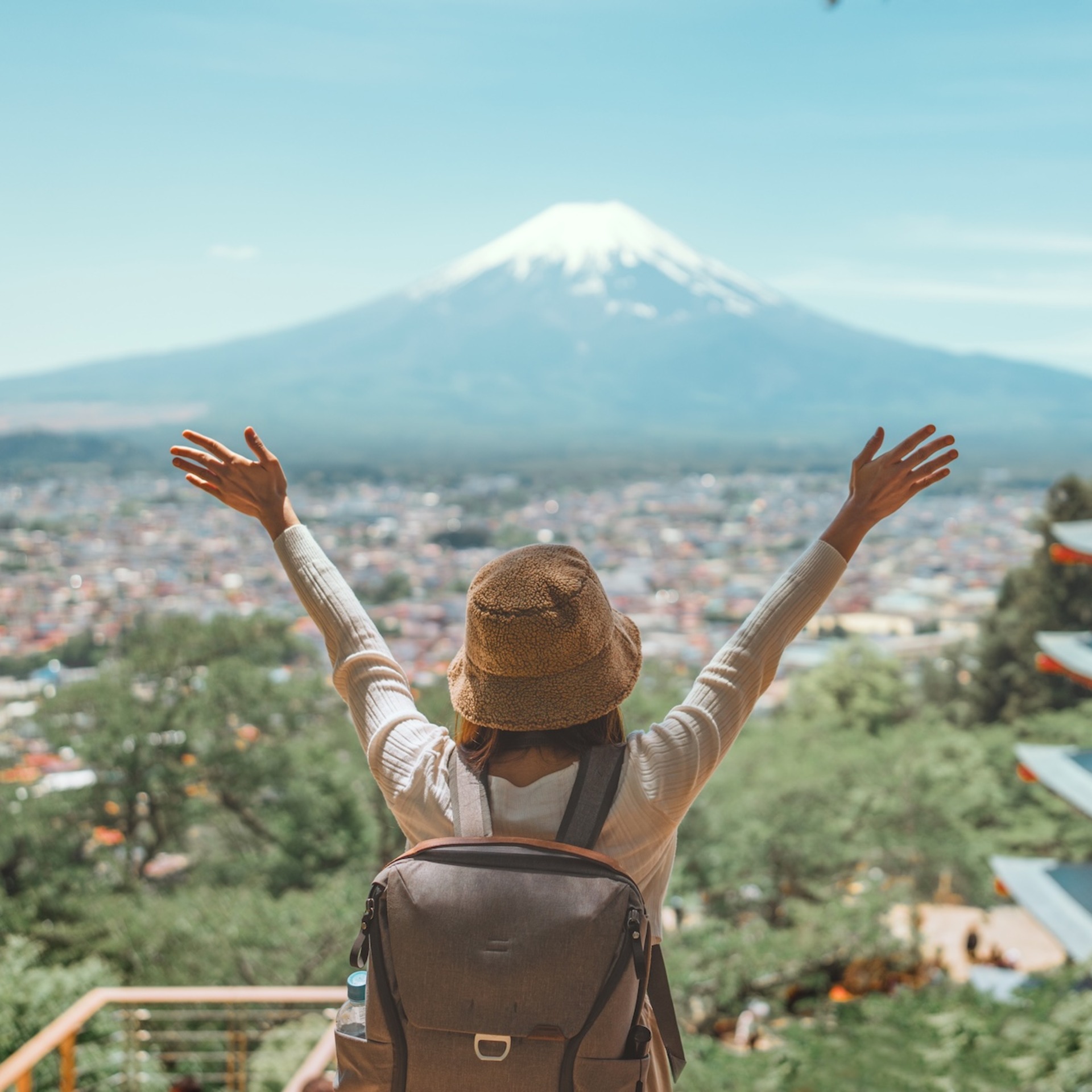 Eine Person mit Hut und Rucksack steht mit ausgestreckten Armen und Rücken zur Kamera in der Natur. In der Ferne ist der Berg Fuji zu sehen mit einer schneebedeckten Spitze und eine Stadt, mit einer Pagode auf der rechten Seite.