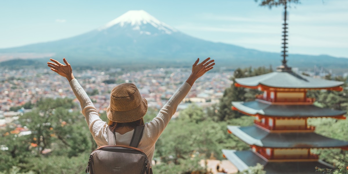 Eine Person mit Hut und Rucksack steht mit ausgestreckten Armen und Rücken zur Kamera in der Natur. In der Ferne ist der Berg Fuji zu sehen mit einer schneebedeckten Spitze und eine Stadt, mit einer Pagode auf der rechten Seite.