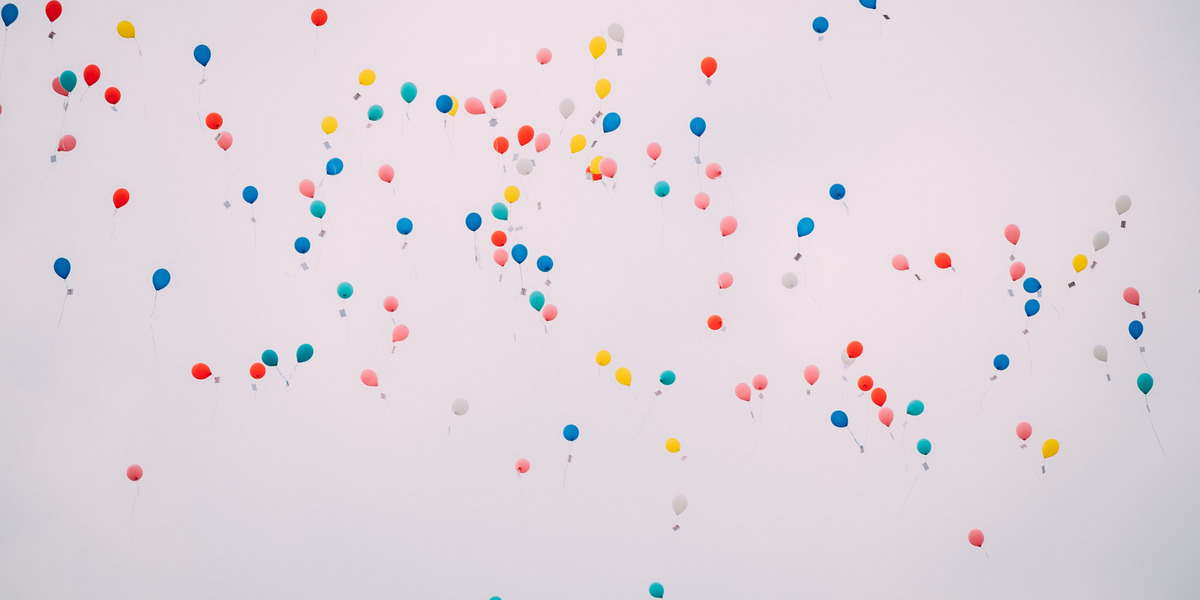 Blick in einen hellgrauen Himmel in dem zahlreiche bunte Luftballons schweben.