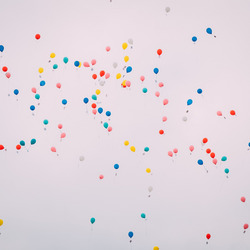 Blick in einen hellgrauen Himmel in dem zahlreiche bunte Luftballons schweben.