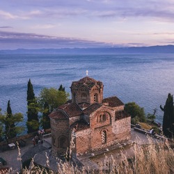 Eine byzantinische Backsteinkirche mit roten Ziegeldächern steht malerisch auf einer Klippe am Ufer eines großen Sees, umgeben von Zypressen im Abendlicht. Im Hintergrund erstreckt sich der See bis zu fernen Bergen unter einem bewölkten Himmel in Rosa- und Blautönen.