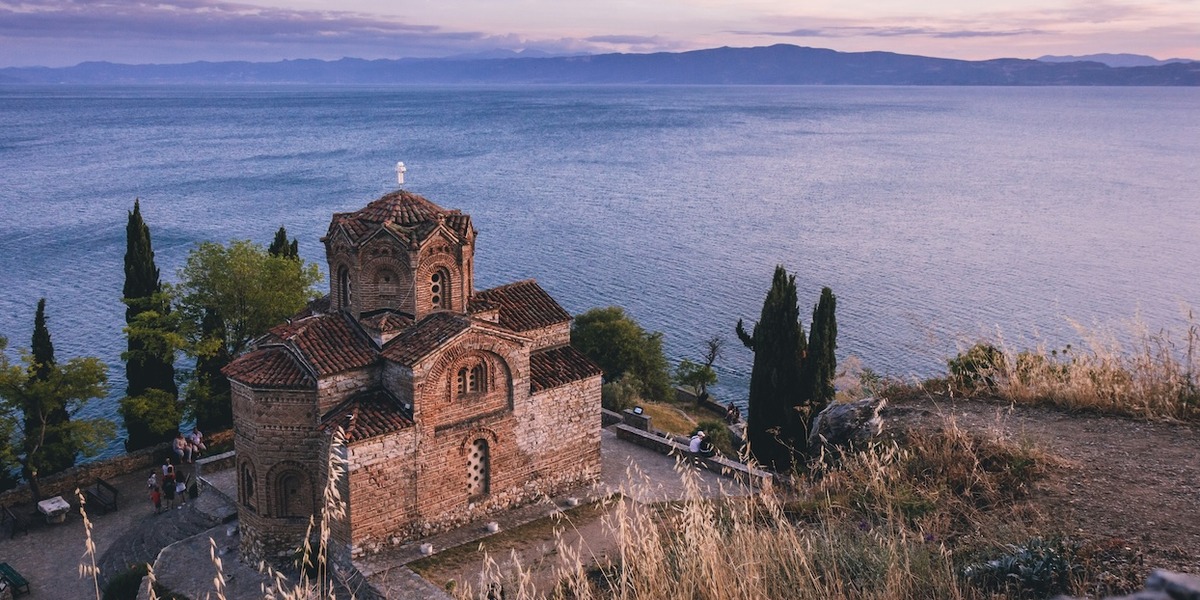 Eine byzantinische Backsteinkirche mit roten Ziegeldächern steht malerisch auf einer Klippe am Ufer eines großen Sees, umgeben von Zypressen im Abendlicht. Im Hintergrund erstreckt sich der See bis zu fernen Bergen unter einem bewölkten Himmel in Rosa- und Blautönen.