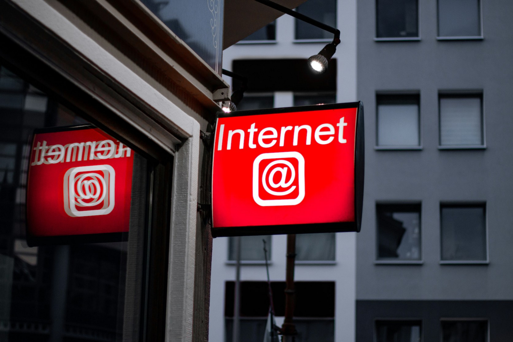 Ein leuchtendes rotes Schild mit der Aufschrift 'Internet' und einem @-Symbol, das an der Außenwand eines Gebäudes angebracht ist. Das Schild wird von einer Lampe angestrahlt und spiegelt sich im Fenster daneben. Im Hintergrund sind unscharfe Wohngebäude zu sehen.