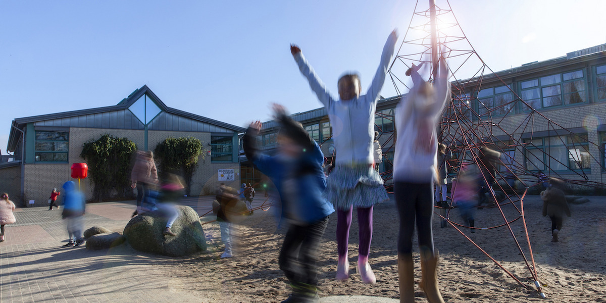 Kinder spielen ausgelassen auf einem Schulhof. Im Vordergrund springen mehrere Kinder mit erhobenen Armen von Felsen, ihre Bewegungen sind verschwommen. Im Hintergrund sieht man ein Schulgebäude mit großen Fenstern und eine rote Kletterstruktur. Die Szene strahlt Energie und Freude aus.