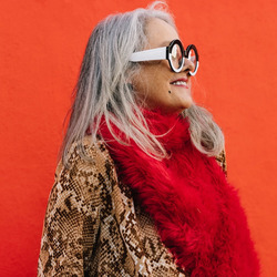 Im Mittelpunkt steht eine ältere Frau mit langen grauen Haaren und runder Brille, die einen roten Schal und eine braun-beige Jacke mit Schlangenmuster trägt. Sie lächelt und schaut zur Seite vor einem leuchtend roten Hintergrund.