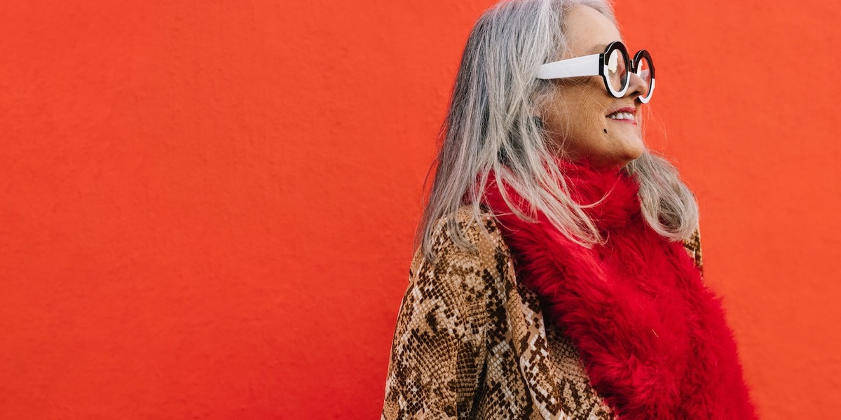 Im Mittelpunkt steht eine ältere Frau mit langen grauen Haaren und runder Brille, die einen roten Schal und eine braun-beige Jacke mit Schlangenmuster trägt. Sie lächelt und schaut zur Seite vor einem leuchtend roten Hintergrund.
