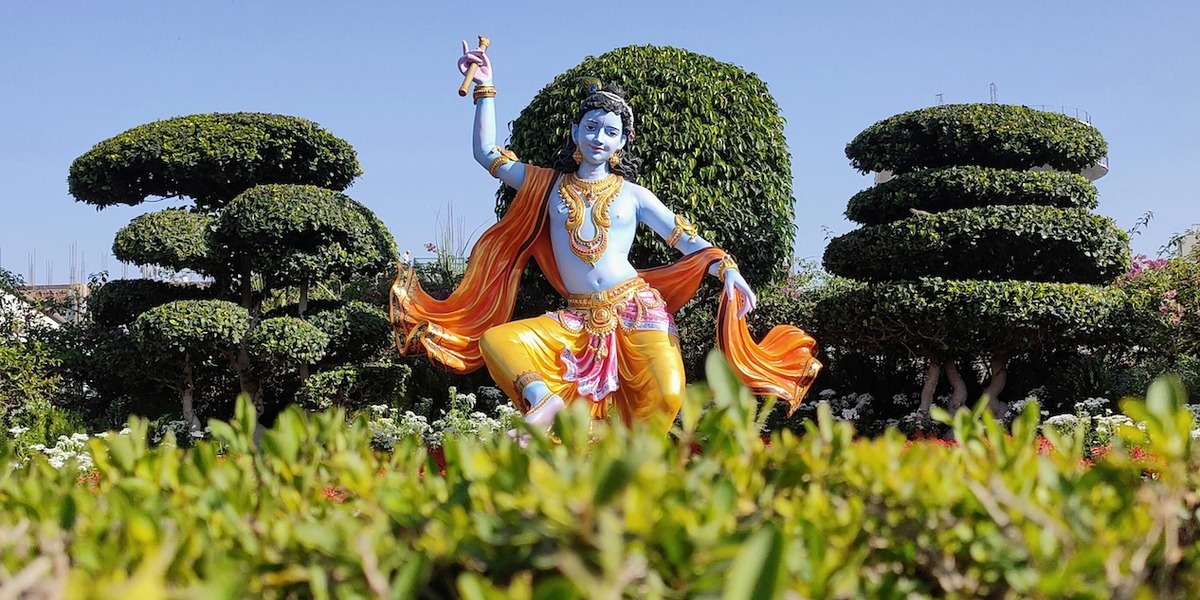 Statue des hinduistischen Gottes Krishna mit blauer Haut, goldgelber Kleidung und orangefarbenem Umhang in einem kunstvoll angelegten Garten mit geschnittenen Bäumen unter blauem Himmel.