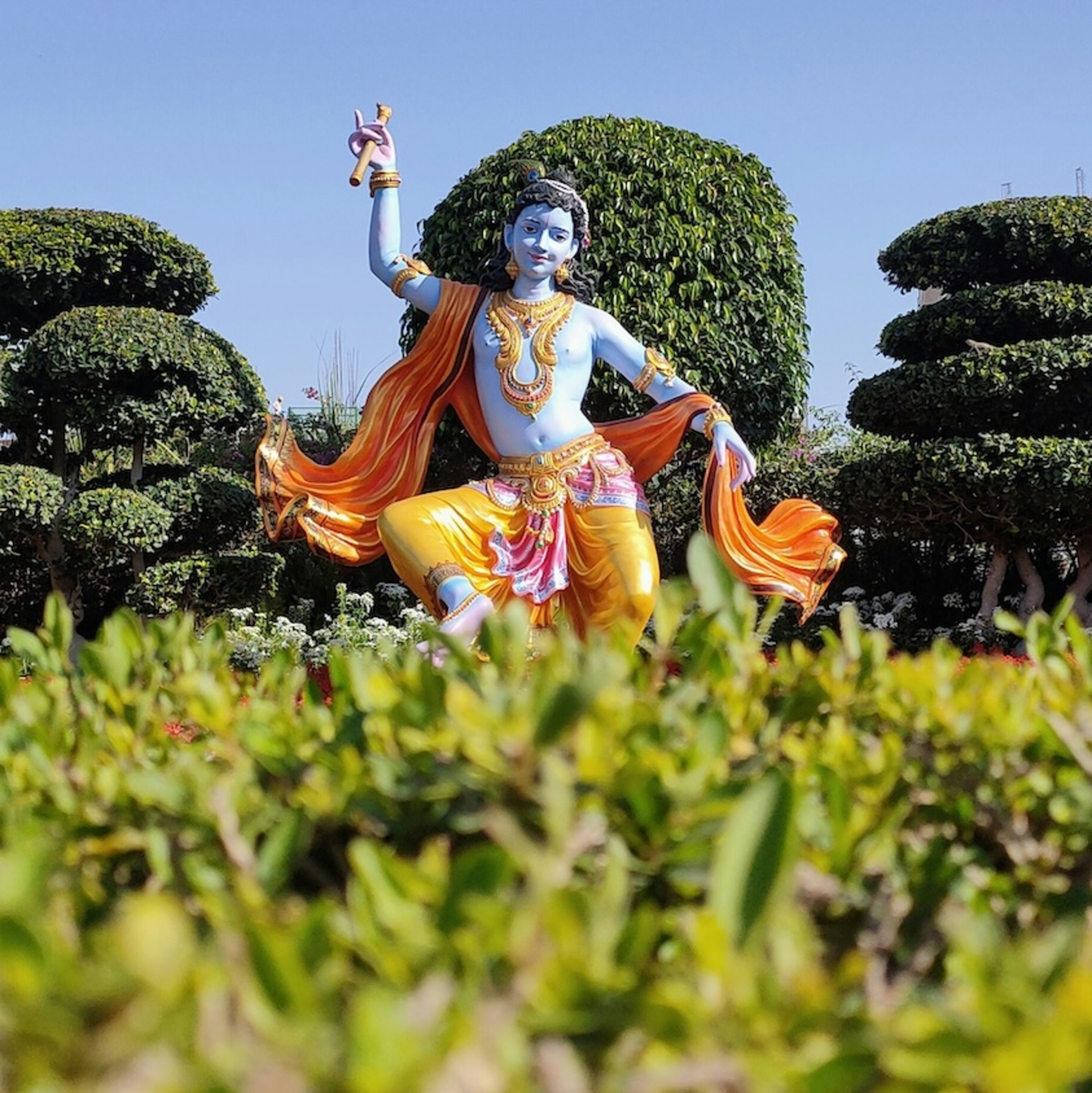 Statue des hinduistischen Gottes Krishna mit blauer Haut, goldgelber Kleidung und orangefarbenem Umhang in einem kunstvoll angelegten Garten mit geschnittenen Bäumen unter blauem Himmel.
