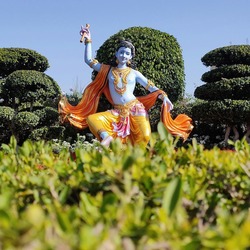 Statue des hinduistischen Gottes Krishna mit blauer Haut, goldgelber Kleidung und orangefarbenem Umhang in einem kunstvoll angelegten Garten mit geschnittenen Bäumen unter blauem Himmel.