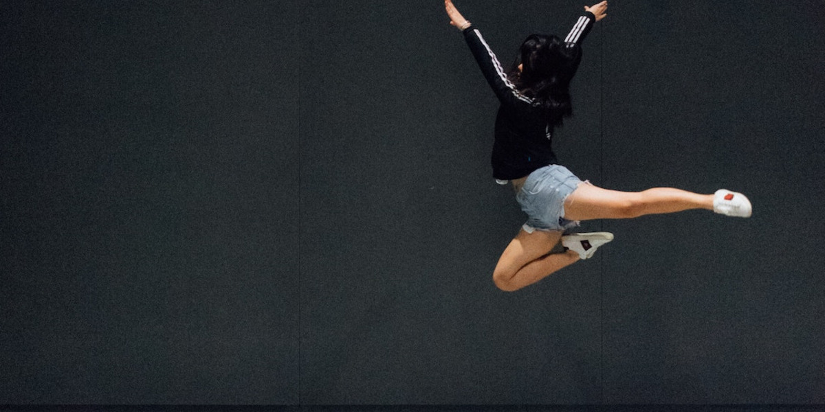 Eine Frau springt in einer Tanzpose vor einer schwarzen Wand hoch.