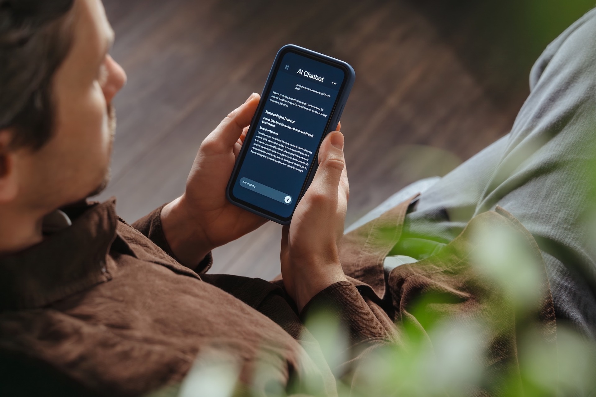 Eine Person sitzt entspannt auf einem Sofa und hält ein Smartphone in beiden Händen. Auf dem Bildschirm ist ein KI-Chatbot geöffnet. Im Vordergrund sind unscharfe grüne Pflanzenblätter zu sehen.