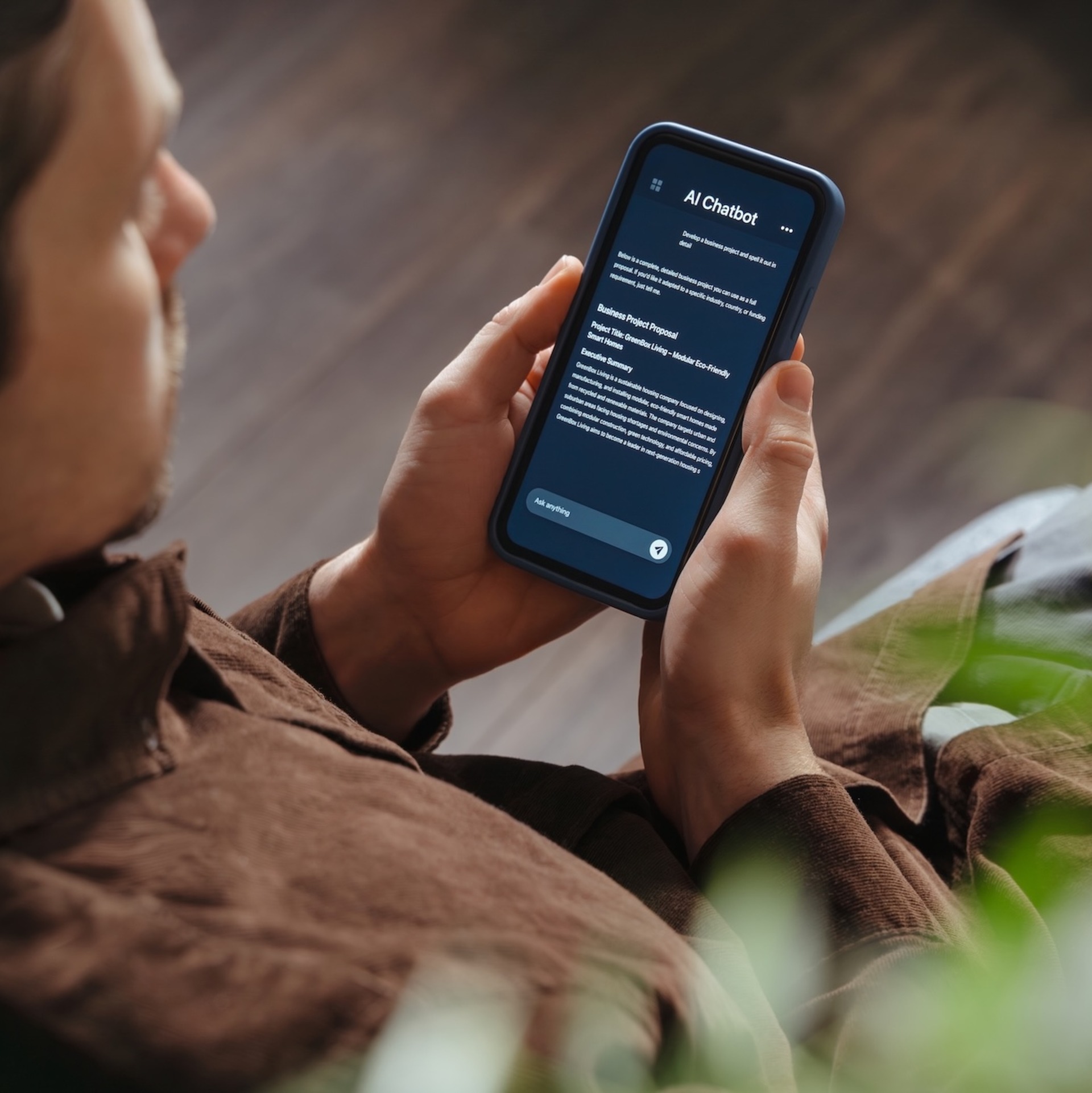 Eine Person sitzt entspannt auf einem Sofa und hält ein Smartphone in beiden Händen. Auf dem Bildschirm ist ein KI-Chatbot geöffnet. Im Vordergrund sind unscharfe grüne Pflanzenblätter zu sehen.