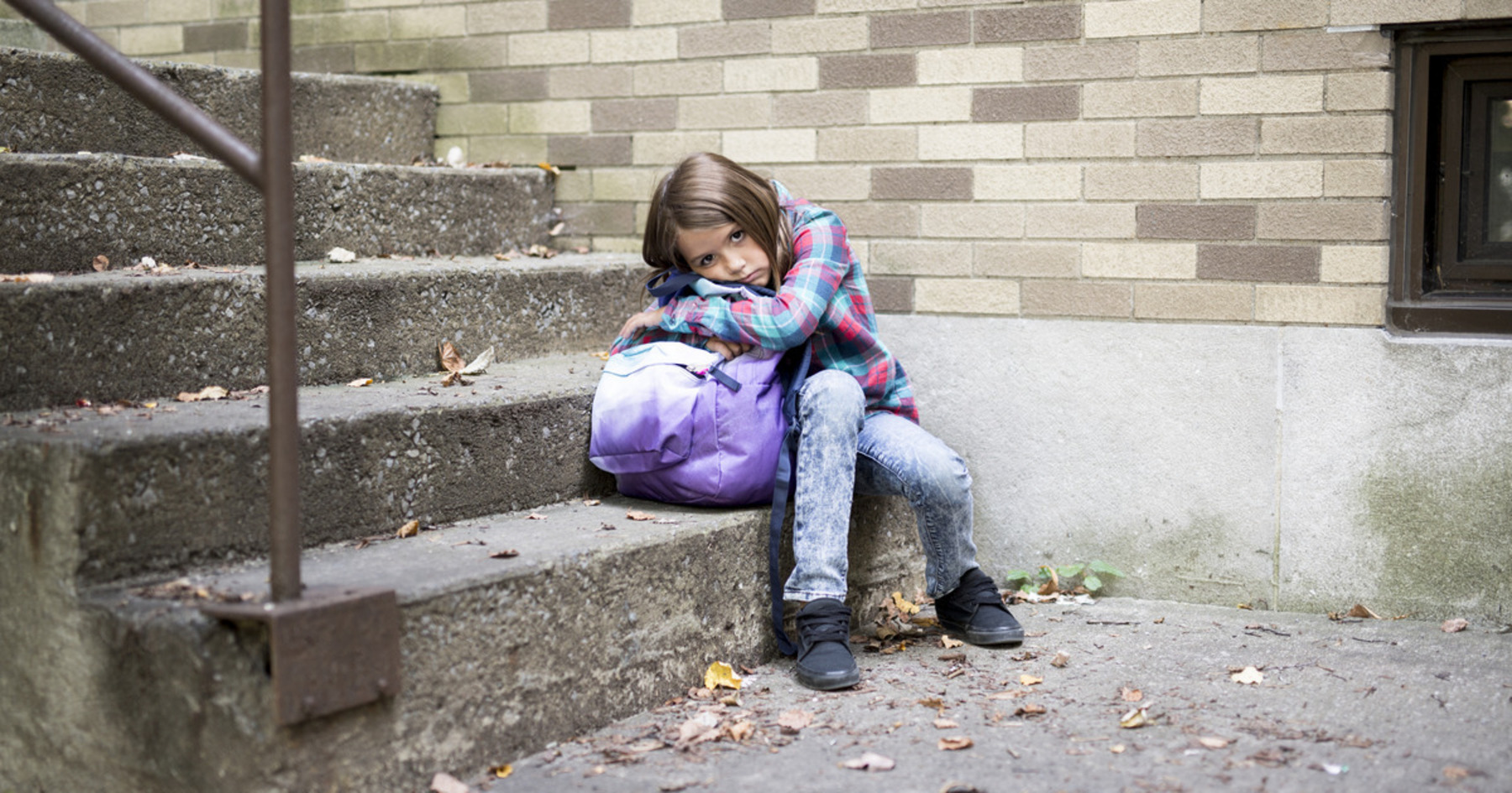 Ein Kind sitzt mit seinem Rucksack auf einer Treppe und sieht etwas traurig aus.