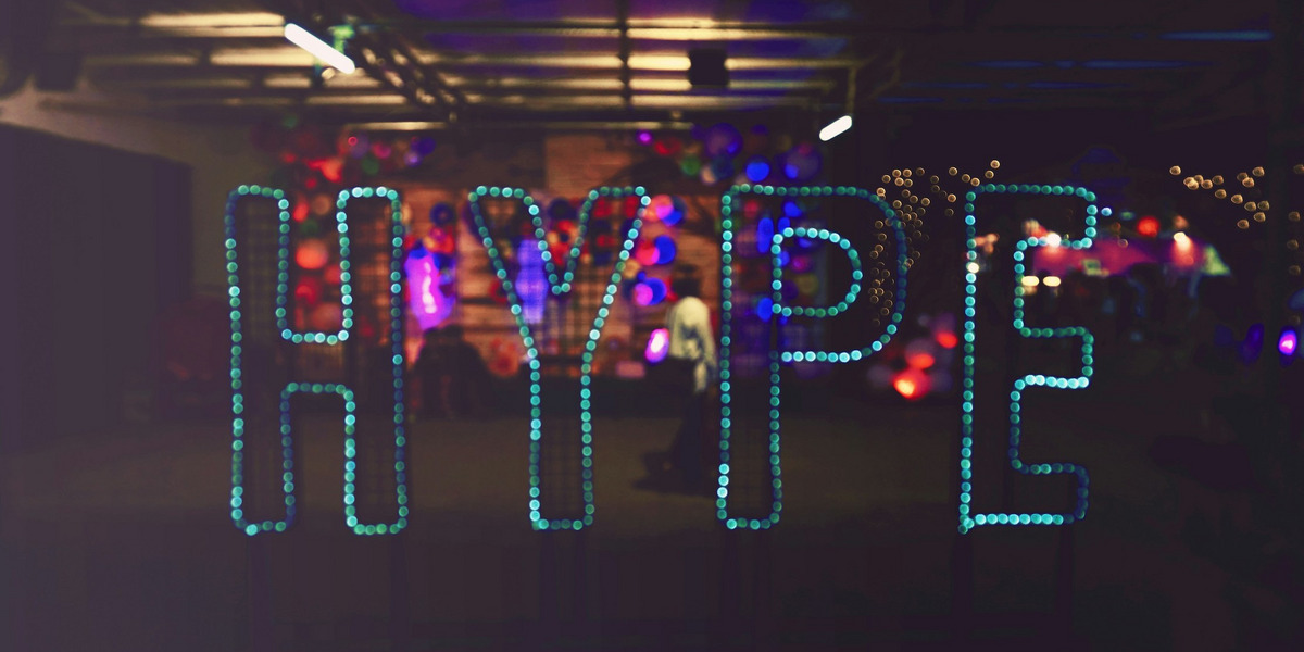 Das Wort 'HYPE' ist aus türkisfarbenen LED-Punkten oder kleinen Leuchten geformt und wird an einer Wand oder im Raum präsentiert. Im Hintergrund sind verschwommene bunte Lichter in Rot, Blau und Violett zu sehen.