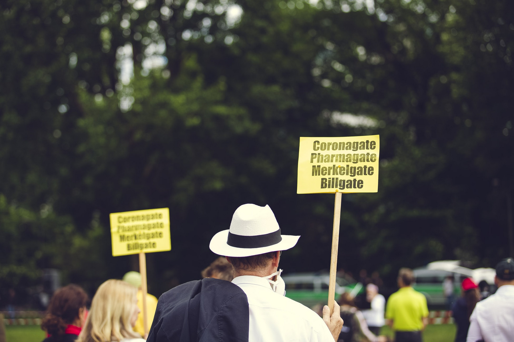 Mann mit Hut hält ein Schild mit der Aufschrift "Coronagate, Pharmagate, Merkelgate, Billgate"