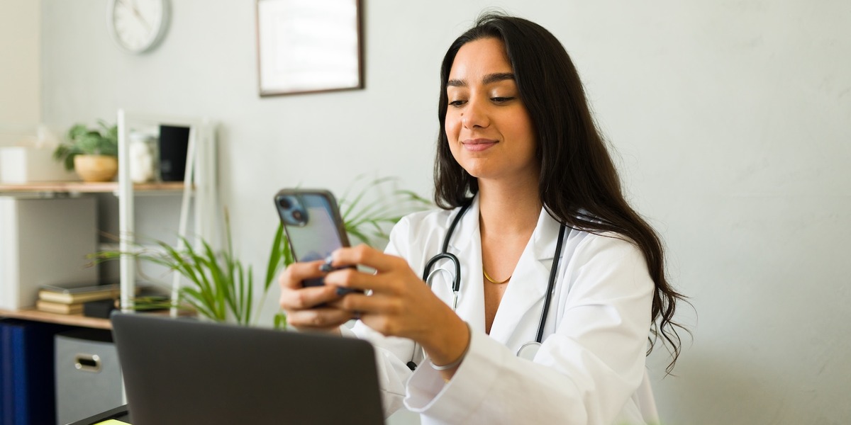 Zu sehen ist eine Ärztin im weißen Kittel mit Stethoskop sitzt an einem Schreibtisch und schaut lächelnd auf ihr Smartphone. Im Hintergrund sind ein Laptop, Regale mit Pflanzen und eine Wanduhr zu sehen.