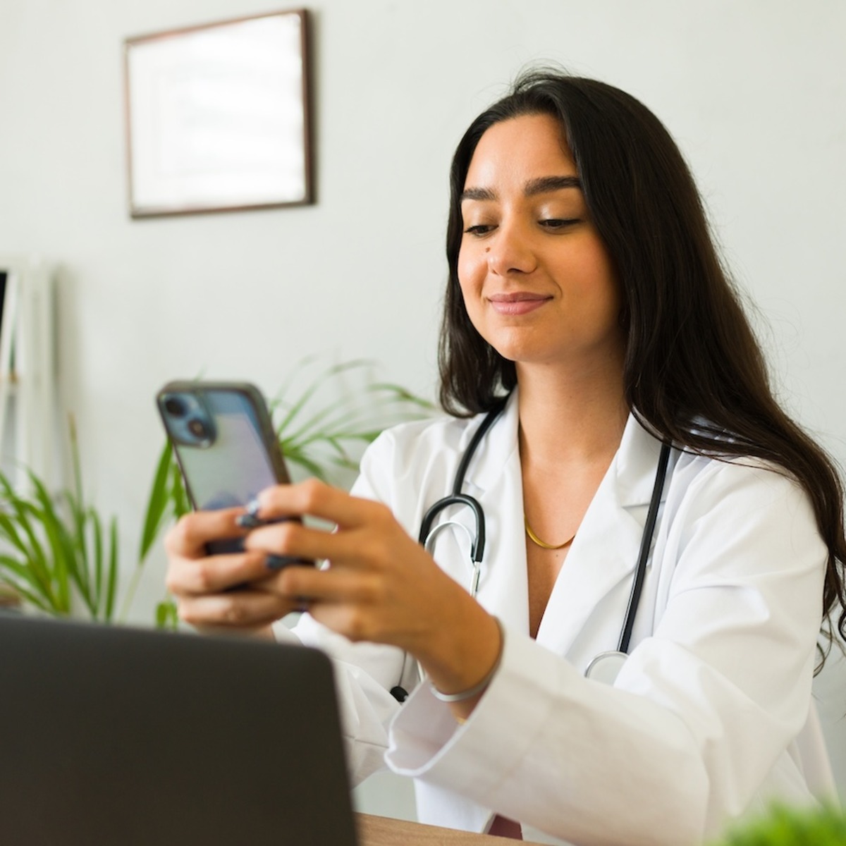 Zu sehen ist eine Ärztin im weißen Kittel mit Stethoskop sitzt an einem Schreibtisch und schaut lächelnd auf ihr Smartphone. Im Hintergrund sind ein Laptop, Regale mit Pflanzen und eine Wanduhr zu sehen.