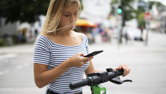 Das Foto zeigt eine junge Frau bei Tageslicht auf der Straße, der Hintergrund ist unscharf, aber man erahnt eine Kreuzung mit einer grünen Ampel in der Ferne. Die Frau hält ein Smartphone in der rechten Hand, ihre linke Hand umgreift den Lenker eines E-Rollers, auf dem sie steht.