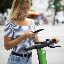 Das Foto zeigt eine junge Frau bei Tageslicht auf der Straße, der Hintergrund ist unscharf, aber man erahnt eine Kreuzung mit einer grünen Ampel in der Ferne. Die Frau hält ein Smartphone in der rechten Hand, ihre linke Hand umgreift den Lenker eines E-Rollers, auf dem sie steht.