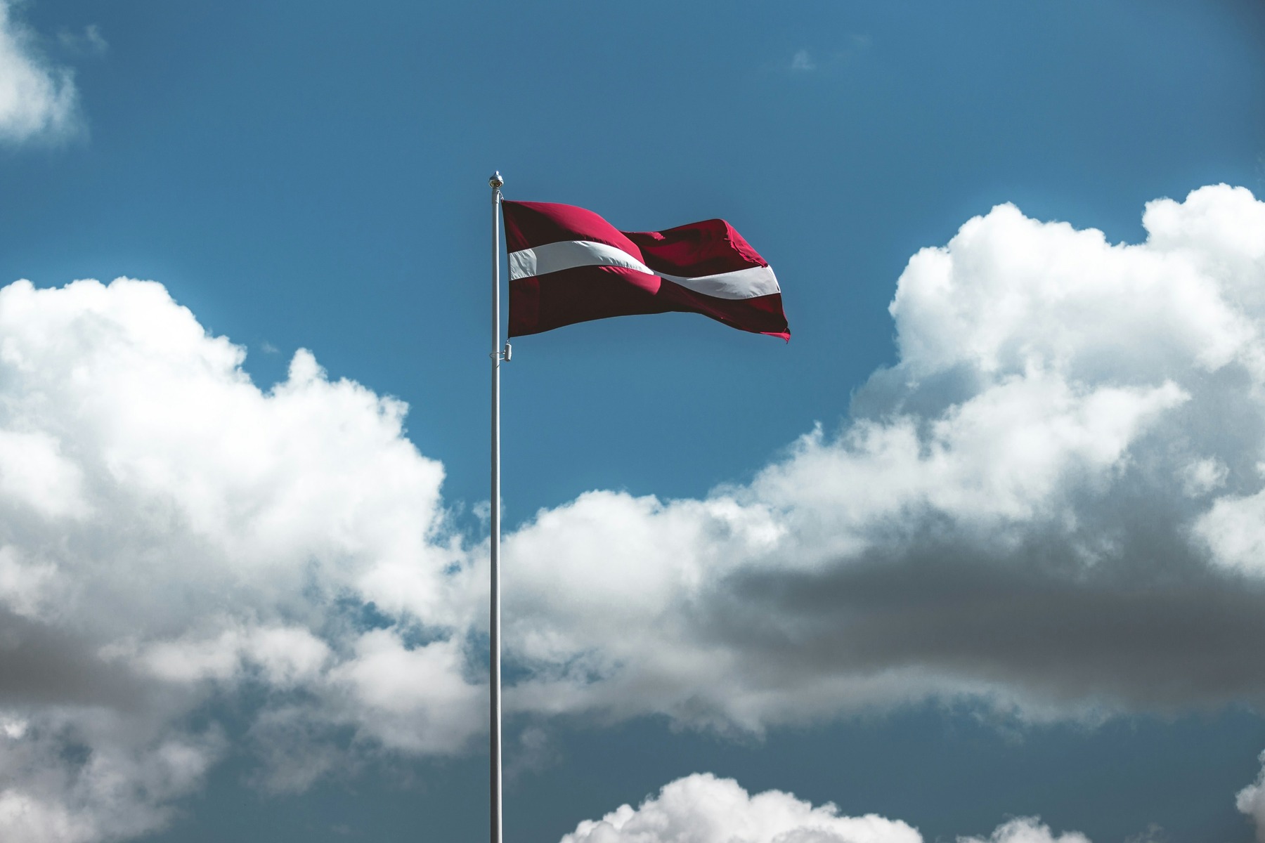 Die lettische Flagge weht an einem hohen Fahnenmast vor einem bewölkten blauen Himmel. Die rot-weiß-rote Fahne ist deutlich gegen die weißen und grauen Wolken zu erkennen.