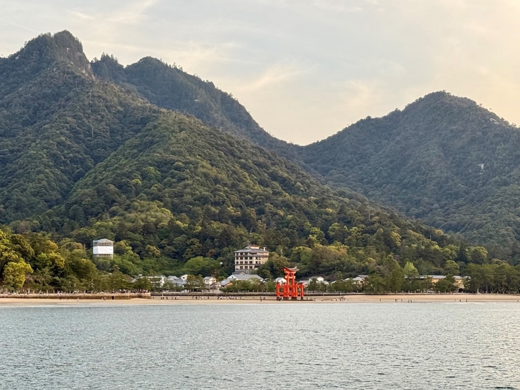 Blick über ruhiges Wasser auf dicht bewaldete Berge mit einem roten Torii-Tor am Ufer und einem Gebäude am Berghang.