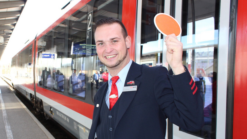 Der Zugbegleiter Manuel Gebhard steht vor einem Zug und schwingt die Kelle auf dem Bahnsteig