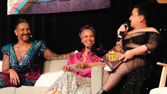 Auf einer Bühne sitzen Oyindamola Alashe und Gianni Jovanovic auf einem Sofa und sprechen mit einer Moderatorin im Rollstuhl, die ein Mikrofon hält. Hinter ihnen hängen Pride-Flaggen und Protestschilder, während sie sich lächelnd unterhalten.