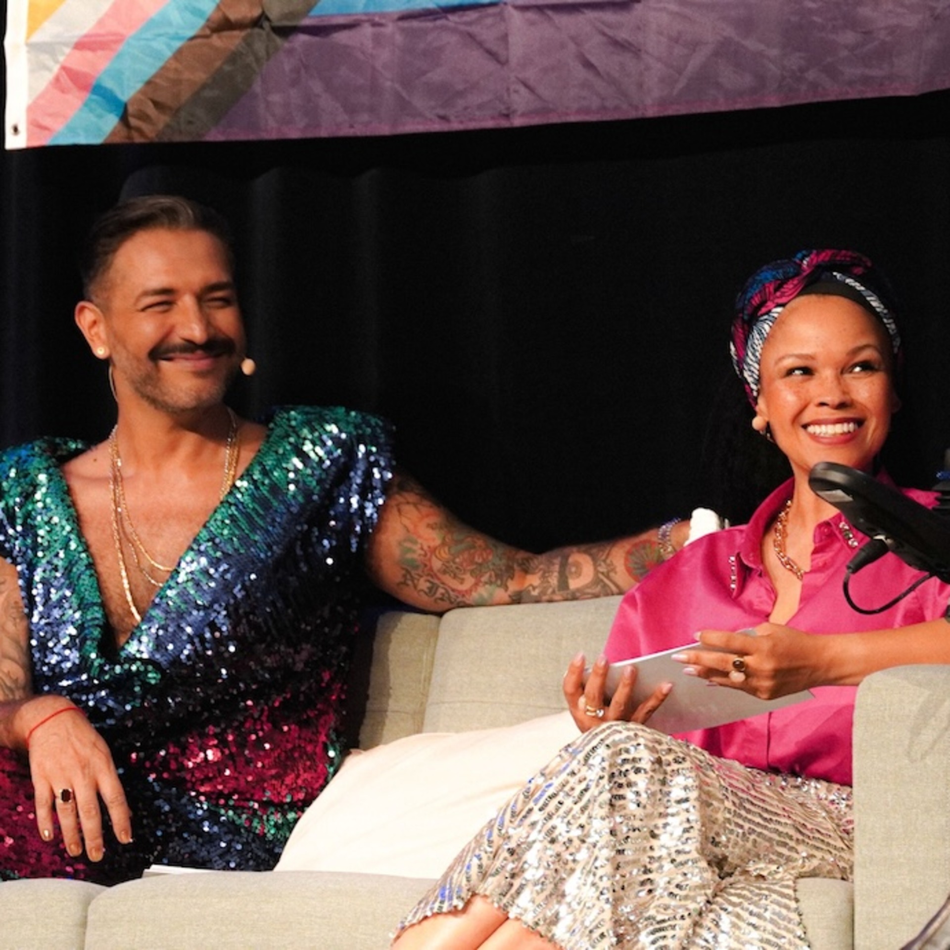 Auf einer Bühne sitzen Oyindamola Alashe und Gianni Jovanovic auf einem Sofa und sprechen mit einer Moderatorin im Rollstuhl, die ein Mikrofon hält. Hinter ihnen hängen Pride-Flaggen und Protestschilder, während sie sich lächelnd unterhalten.