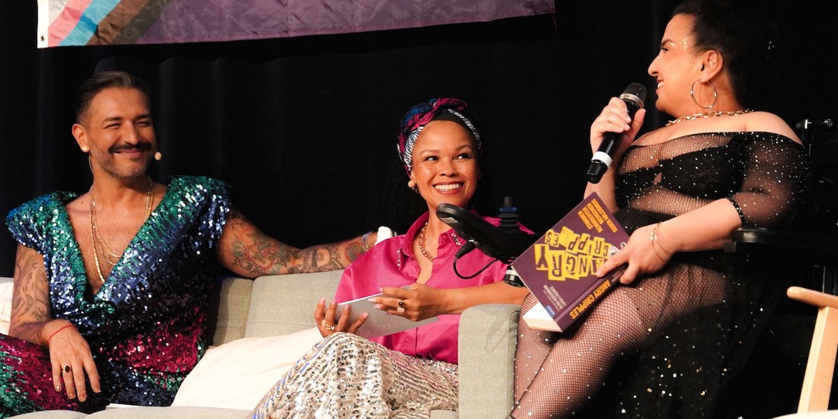 Auf einer Bühne sitzen Oyindamola Alashe und Gianni Jovanovic auf einem Sofa und sprechen mit einer Moderatorin im Rollstuhl, die ein Mikrofon hält. Hinter ihnen hängen Pride-Flaggen und Protestschilder, während sie sich lächelnd unterhalten.