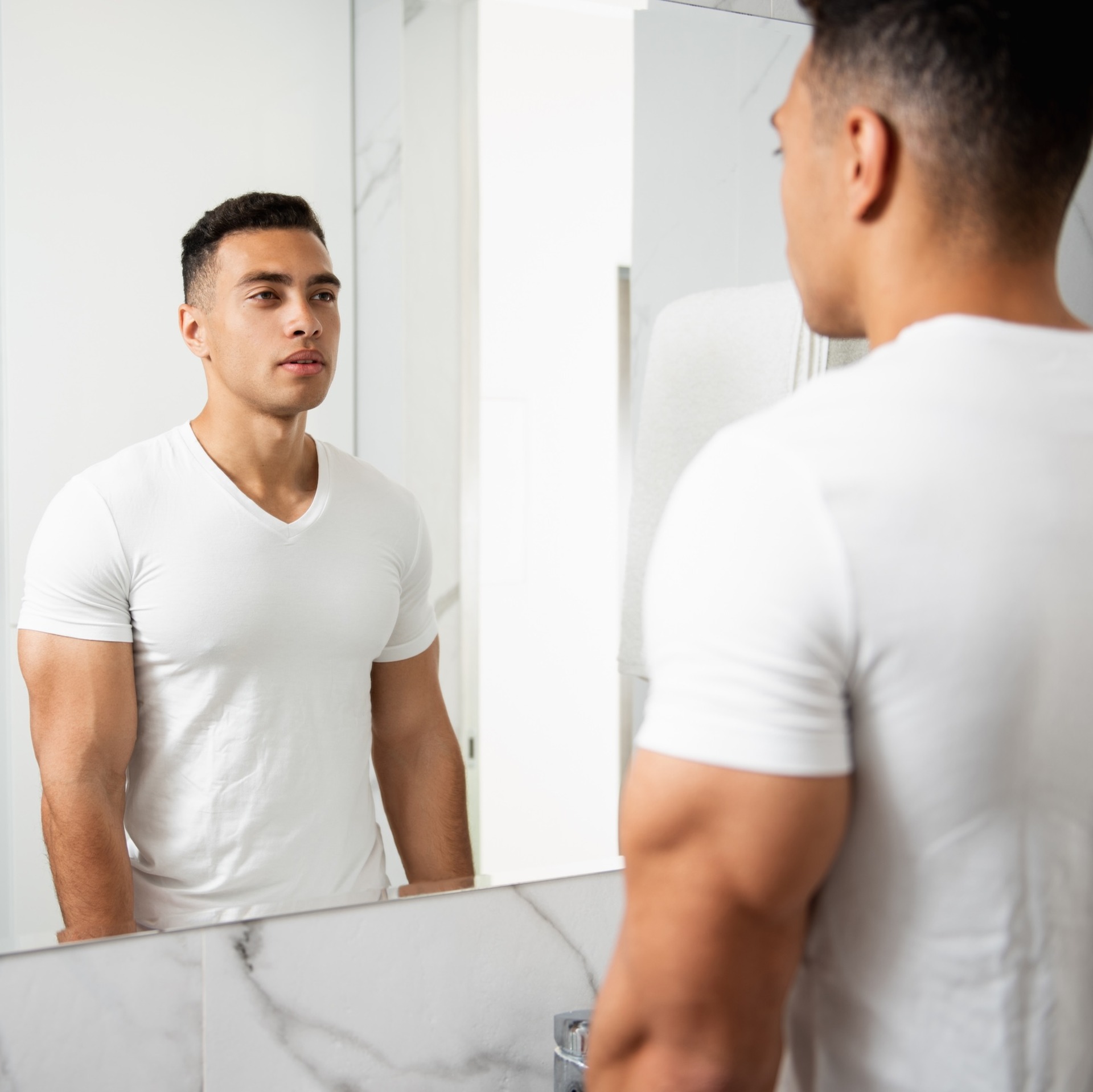 Eine Person steht in einem hell gefliesten Badezimmer vor einem Spiegel. Die Person hat kurze Haare, trägt ein weißes T-Shirt und blickt sich selbst über das Spiegelbild an. Der Oberkörper ist angespannt und muskulös.