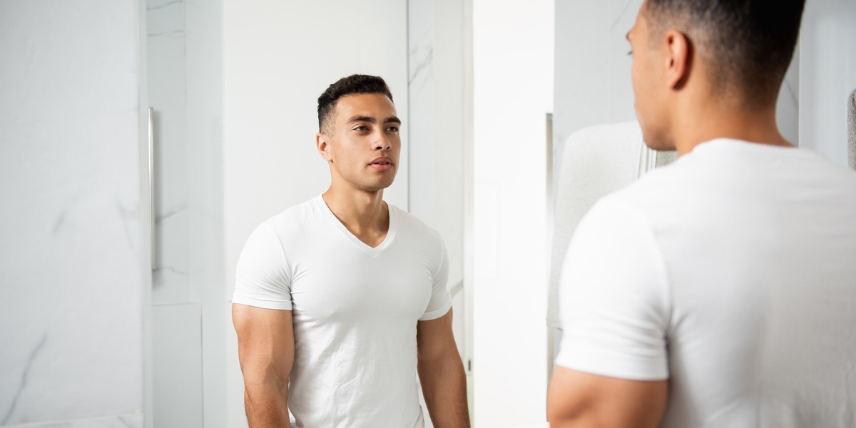 Eine Person steht in einem hell gefliesten Badezimmer vor einem Spiegel. Die Person hat kurze Haare, trägt ein weißes T-Shirt und blickt sich selbst über das Spiegelbild an. Der Oberkörper ist angespannt und muskulös.