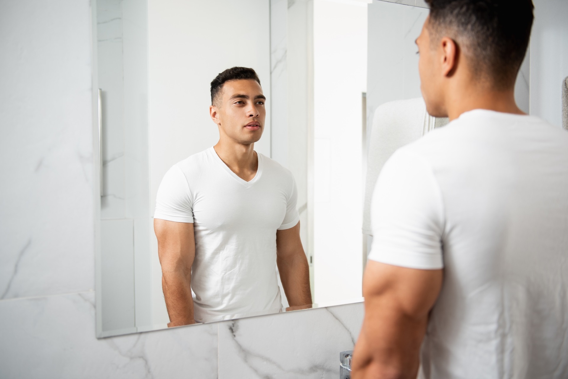 Eine Person steht in einem hell gefliesten Badezimmer vor einem Spiegel. Die Person hat kurze Haare, trägt ein weißes T-Shirt und blickt sich selbst über das Spiegelbild an. Der Oberkörper ist angespannt und muskulös.