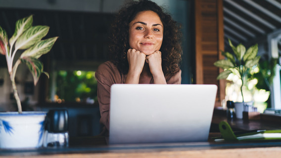 Eine junge Frau mit braunen Locken sitzt vor einem Laptop und hat das Kinn auf die Hände gestützt.