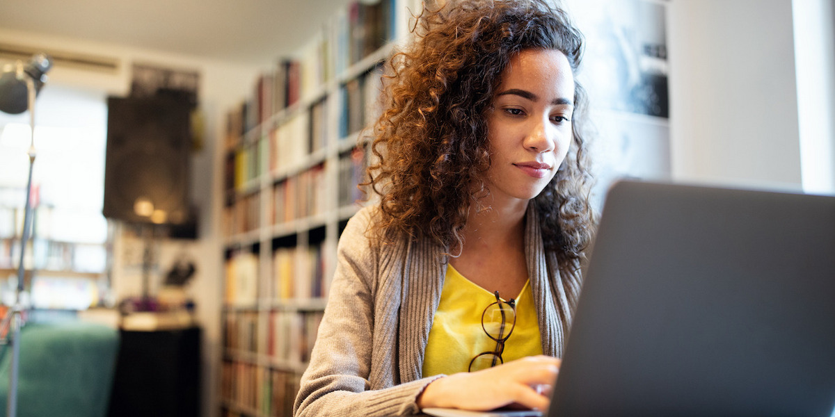 Junge Frau mit langen braunen Haaren sitzt lächelnd am Laptop. Sie trägt ein gelbes T-Shirt und eine graue Jacke. Im Hintergrund sind Bücherregale zu sehen.