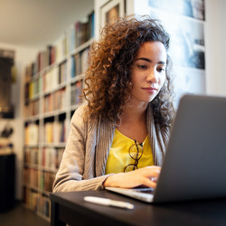 Junge Frau mit langen braunen Haaren sitzt lächelnd am Laptop. Sie trägt ein gelbes T-Shirt und eine graue Jacke. Im Hintergrund sind Bücherregale zu sehen.
