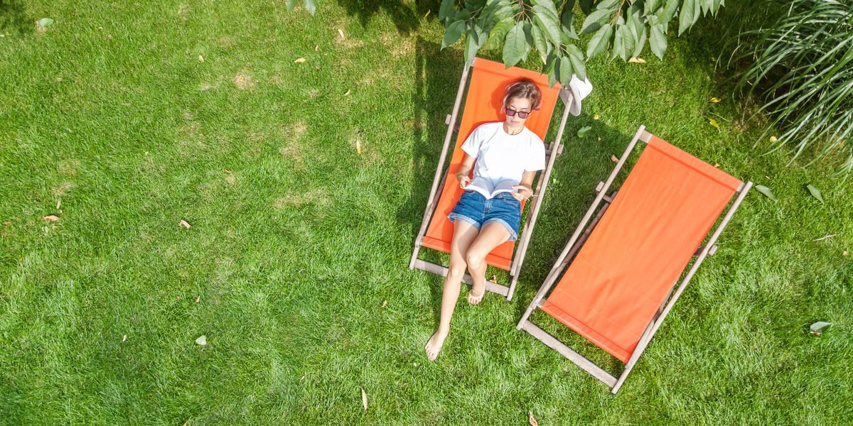 Frau liegt auf einer orangefarbenen Sonnenliege im Garten und liest ein Buch. Neben ihr steht eine zweite, leere Sonnenliege auf der grünen Wiese.