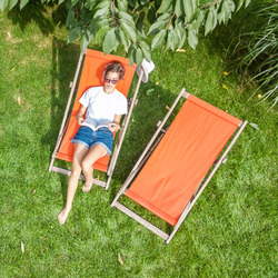 Frau liegt auf einer orangefarbenen Sonnenliege im Garten und liest ein Buch. Neben ihr steht eine zweite, leere Sonnenliege auf der grünen Wiese.