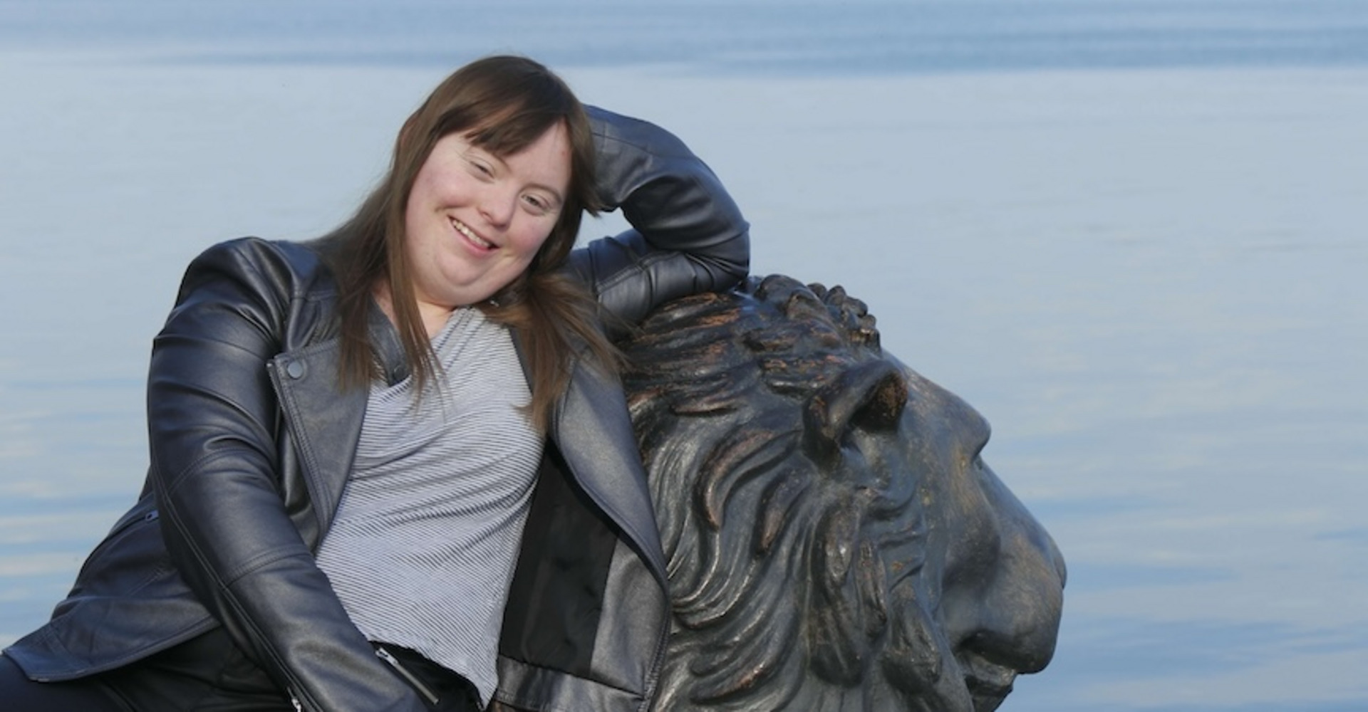 Luisa Wöllisch, eine Frau mit langen braunen Haaren und einem grauen Lederjackett, lehnt sich entspannt an eine Löwenstatue. Im Hintergrund sieht man einen ruhigen See und eine bewaldete Uferlinie. Luisa lächelt und wirkt glücklich.